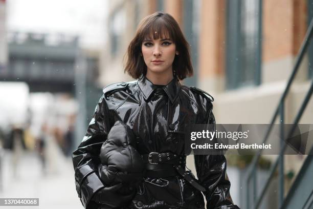 Hari Nef wears a black high neck pullover, a black shiny vinyl belted long trench coat, a black matte puffy leather handbag from Bottega Veneta, a...