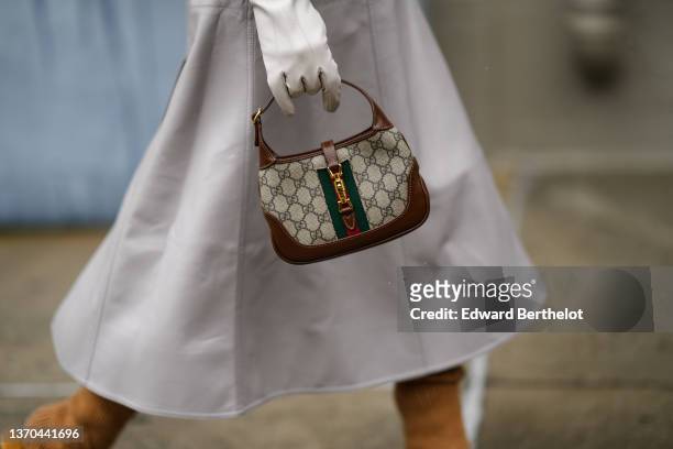Mary Leest wears a pale gray shiny leather long belted trench coat, white leather gloves, a brown leather with beige and gray GG monogram print...