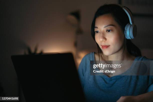 concentrated young asian woman wearing headphones, working and studying online from home on laptop in bedroom till late night. woman using laptop at home, device screen light illuminated on her. working from home, e-learning, hot desking concept - chinese student laptop stock pictures, royalty-free photos & images
