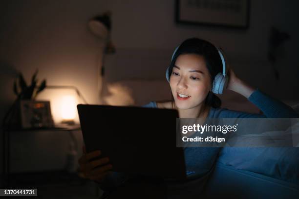 relaxed young asian woman wearing headphones watching a movie on laptop at late night at cozy bedroom. woman using laptop at home, device screen light illuminated on her. lifestyle, habits and technology - asian watching tv stock pictures, royalty-free photos & images