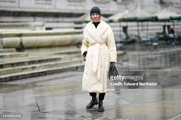 Lisa Aiken wears a dark gray beanie, a black turtleneck pullover, a pale yellow / white latte belted oversized puffer jacket, black suit pants, a...