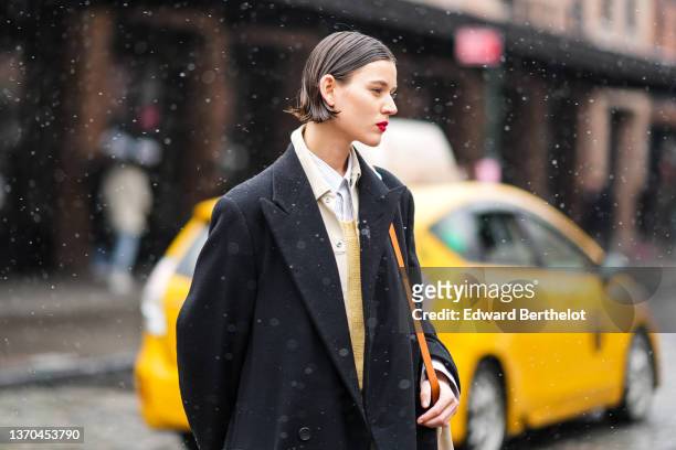 Model wears a white striped print pattern shirt, a pale yellow wool pullover, a beige shirt jacket, a black long coat, an orange shiny leather and...
