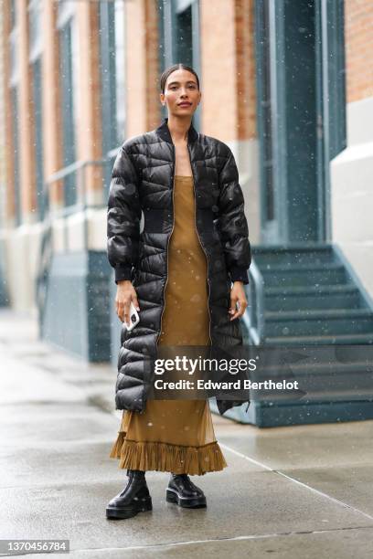 Guest wears silver earrings, a brown square-neck long dress with ruffled pattern ankle, a black shiny belted long puffer jacket, a silver ring, black...