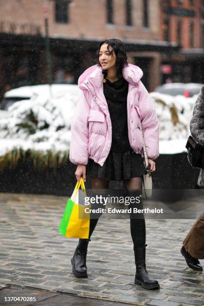 Model wears a black high neck pullover, a black short pleated skirt, a pale pink hoodie oversized puffer jacket, black tights, black stockings / high...
