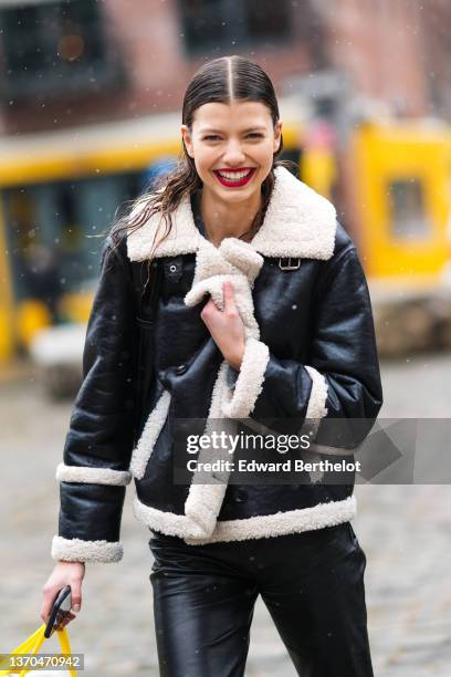 Model wears a gray wool pullover, a black shiny leather with white sheep interior aviator coat, black shiny leather cigarette pants, a green and...