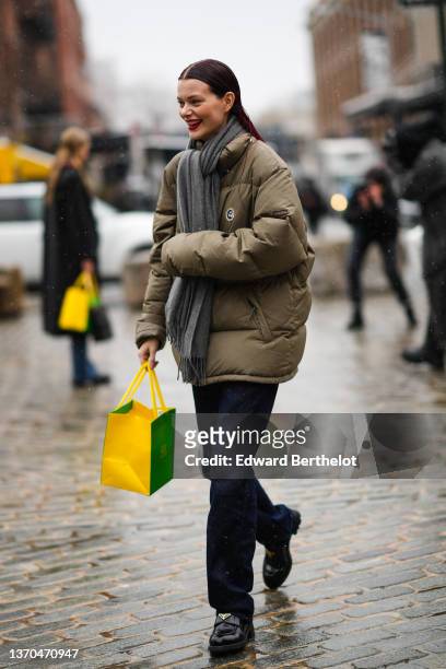 Model wears a grey fringed scarf, a khaki oversized high neck puffer jacket, a green and yellow shopping bag, navy blue denim large jeans pants,...