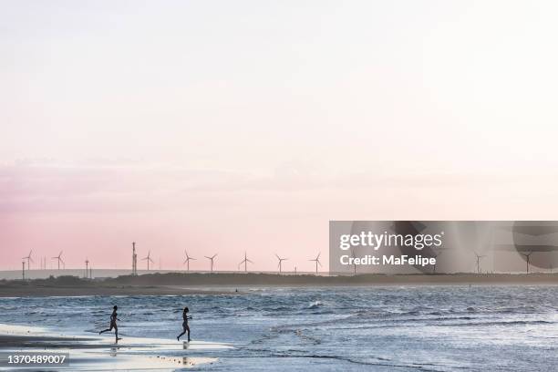 two girls running into the sea in galinhos beach at dusk. many wind turbines for wind power generation are in background. rio grande do norte, northeastern brazil. - brazil ocean stock pictures, royalty-free photos & images