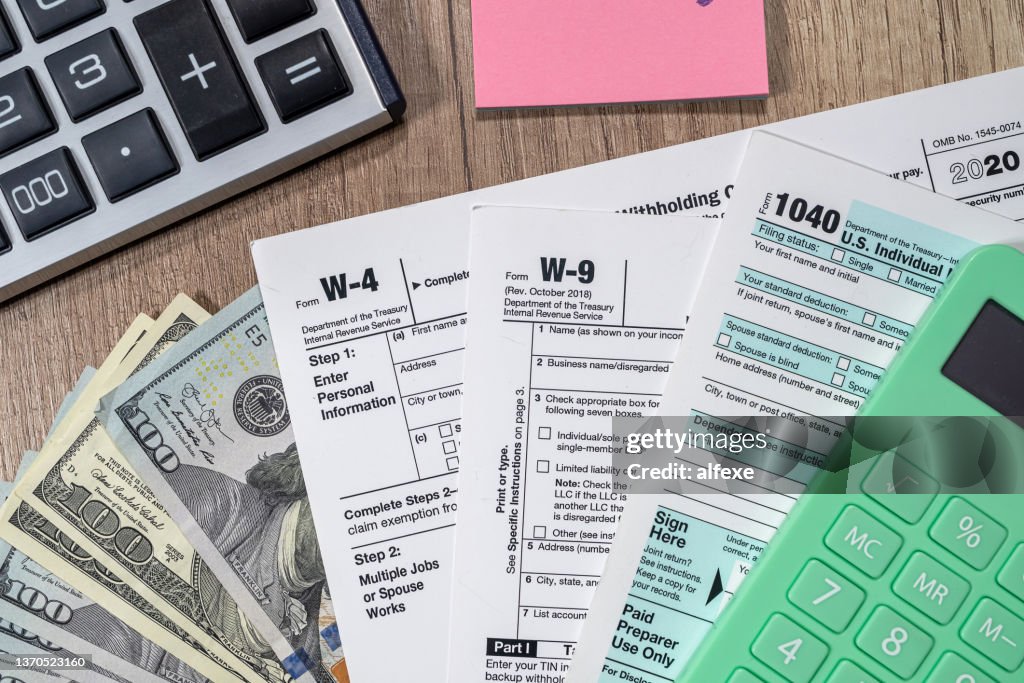 Tax forms 1040 are blank to fill out and dollars are sloppily scattered on a wooden table.