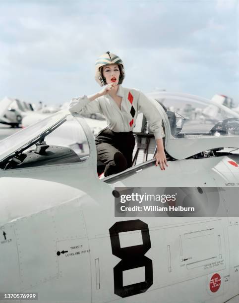 American model and actress Dovima , in a flight helmet and white shirt with red and black diamonds, stands in the cockpit of an F86 Sabre...