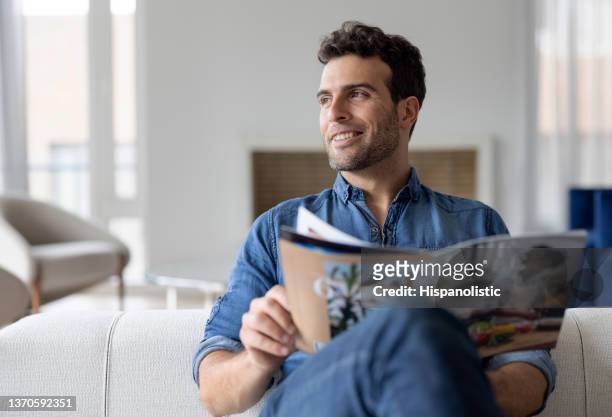 mann zu hause liest eine zeitschrift im wohnzimmer - magazijn stock-fotos und bilder