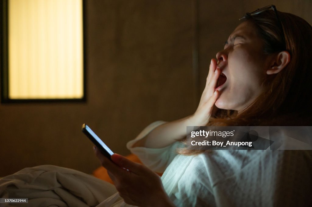 Woman yawning while playing and surfing internet on her smartphone late at night on bed.