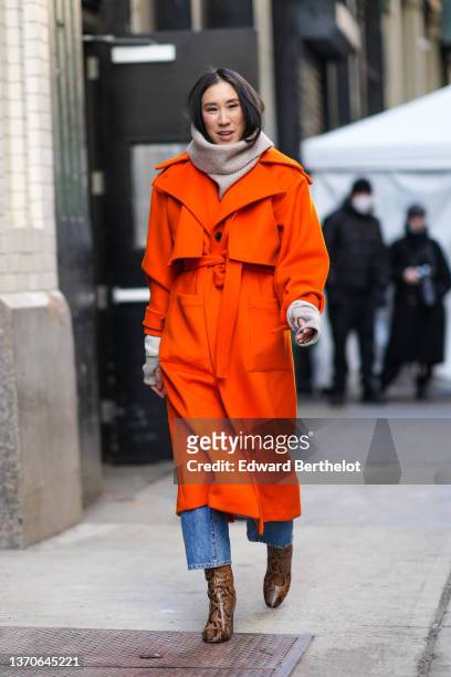Eva Chen wears a beige oversized ribbed wool pullover, a neon orange belted long winter coat, blue denim jeans pants, brown and black snake print...