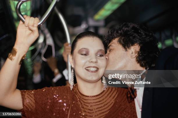 Maria Burton, wearing a red outfit with drop earrings, as her husband, Steve Carson, who wears a tuxedo and bow tie, kisses her as she hangs onto the...