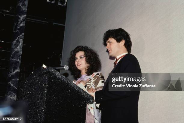 Maria Burton, wearing a lilac, black and peach outfit, and her husband, Steve Carson, who wears a tuxedo, stand behind a lectern as they address an...