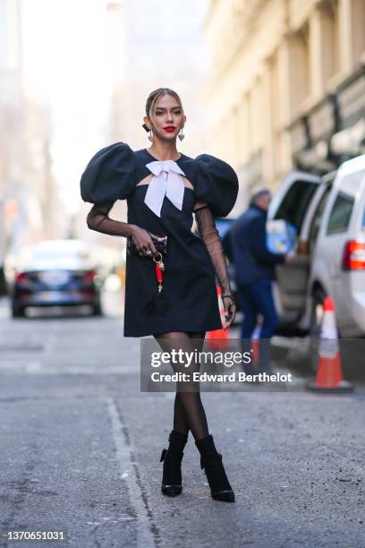 Jordanna Maia wears silver and pearls heart pendant earrings, a black ribbon in the hair, a black tweed oversized puffy shoulder / cut-out chest /...