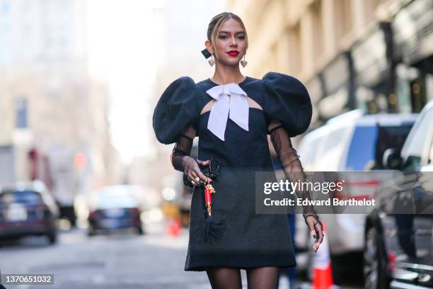 Jordanna Maia wears silver and pearls heart pendant earrings, a black ribbon in the hair, a black tweed oversized puffy shoulder / cut-out chest /...