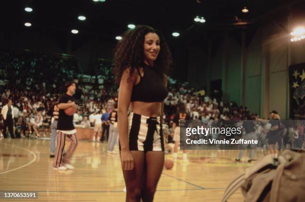 British actress and television personality Downtown Julie Brown, wearing a black crop top with black-and-white striped shorts, courtside during an...