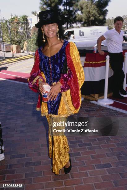 British actress and television personality Downtown Julie Brown, wearing a yellow, blue and red outfit with a black hat, holding a Pepsi cup, circa...
