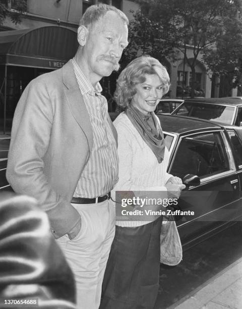 American actress Ellen Burstyn, wearing a white sweater over a cowl neck blouse, accompanied by a man, a motor card in the background, circa 1985.