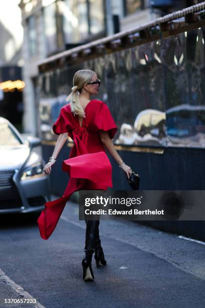 Leonie Hanne wears black sunglasses, gold earrings, a red oversized short sleeves / belted short dress with a large knot train in the back, gold...