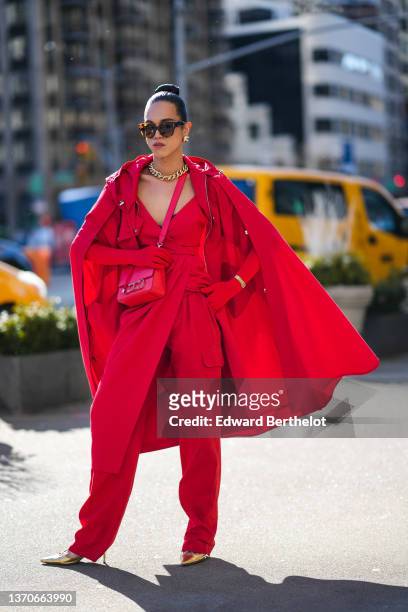 Guest wears black sunglasses from Carolina Herrera, gold large earrings, a gold large chain necklace, a red V-neck wrap slit midi dress, a red...