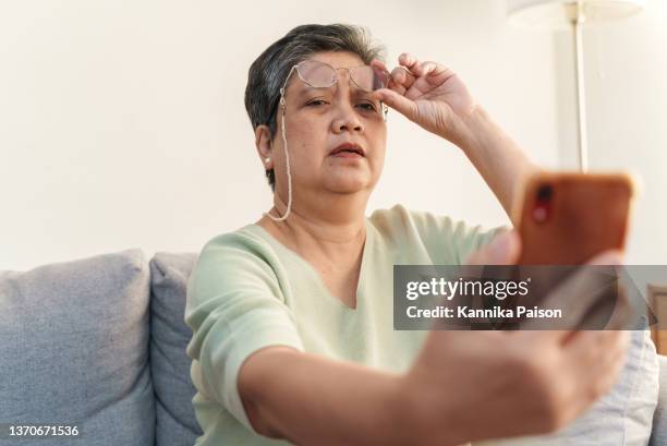 femme asiatique âgée atteinte de presbytie, enlevant des lunettes tout en lisant un message de téléphone intelligent. - effort photos et images de collection