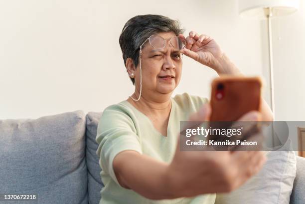 senior asian woman with presbyopia, taking off eyeglasses while reading message from smart phone. - human eye stockfoto's en -beelden