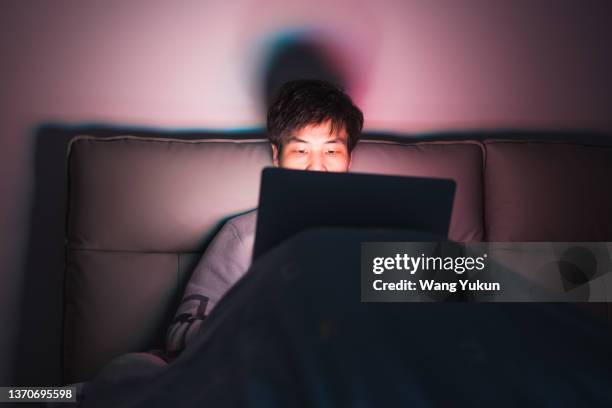 a young asian male sitting on the bed and staying up late for work - staying indoors stock pictures, royalty-free photos & images