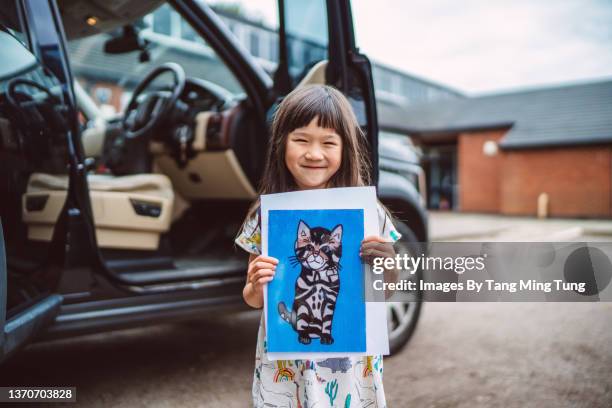 lovely little girl standing next to a car, joyfully showing her drawing of a cat to the camera - kinder lernen spaß stock-fotos und bilder