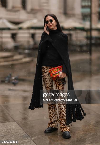 Guest wears a black oversized fringed long scarf, a beige / brown / black leopard print pattern flared pants, black shiny leather ankle boots,...