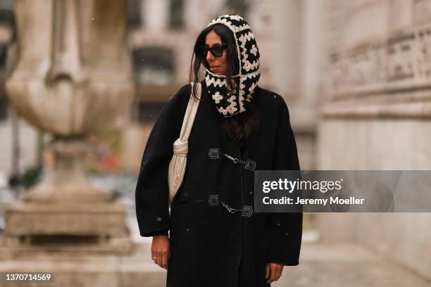 Guest wears a black and white oversized print pattern balaclava, black sunglasses, a black long winter coat, a white latte embossed striped shoulder...