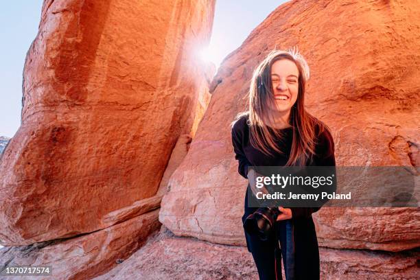 wide angle portrait of a cheerful young woman standing, leaning forward, laughing and holding a professional dslr camera outdoors while visiting and hiking in the colorado national monument - american influencer stock pictures, royalty-free photos & images