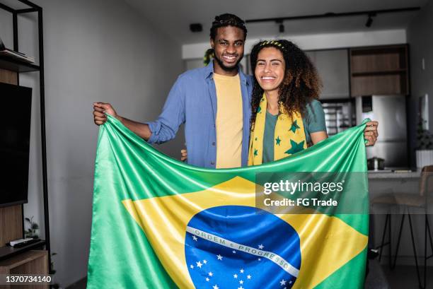 portrait of a happy young couple holding brazilian flag at home - brazil flag stock pictures, royalty-free photos & images