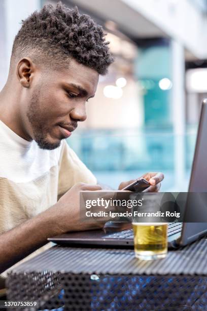 young smiling african american man sitting in a cafe with his laptop sending a message on his mobile. - cafe pink silver stock pictures, royalty-free photos & images