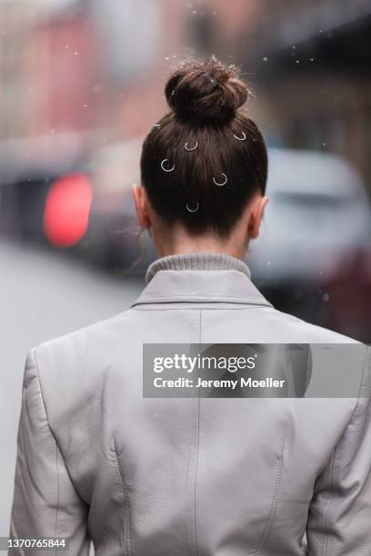 Mary Leest wears silver earrings, a gray ribbed turtleneck pullover, a pale gray shiny leather long belted trench coat, white leather gloves, a brown...