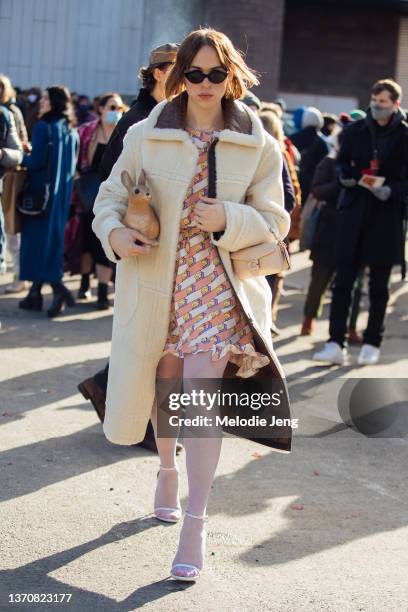 Tommy Dorfman wears a long white shearling jacket, pink patterned baby doll dress, small pink Coach bag tights, white heels, and carries a small...