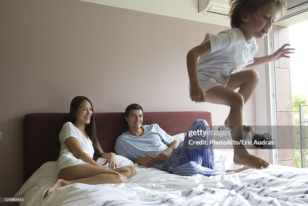 Parents watching as young son jumps on bed