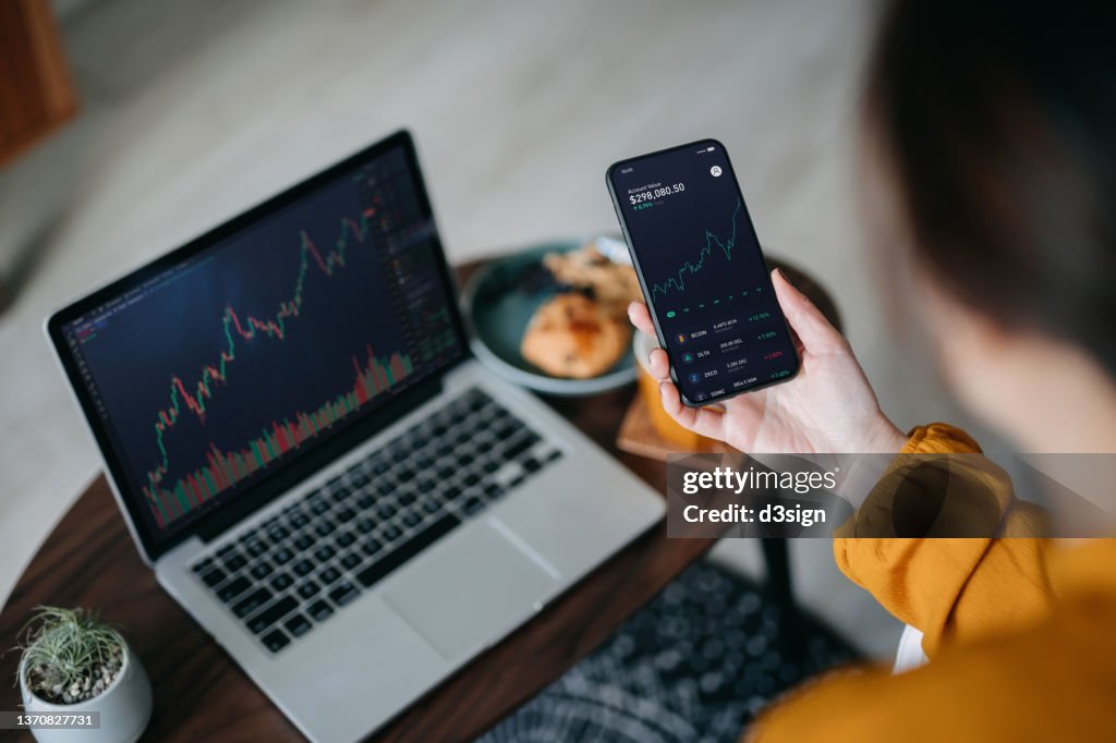Over the shoulder view of Asian woman holding smartphone, analyzing investment trading data on crypto graph with smartphone and laptop while working at home. Cryptocurrency, bitcoin, digital ledger