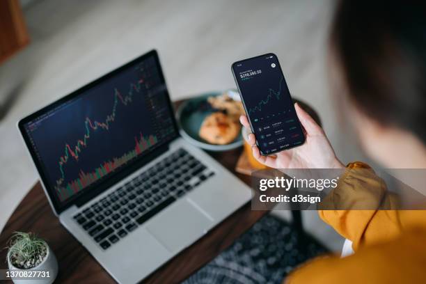 over the shoulder view of asian woman holding smartphone, analyzing investment trading data on crypto graph with smartphone and laptop while working at home. cryptocurrency, bitcoin, digital ledger - woman laptop screen foto e immagini stock