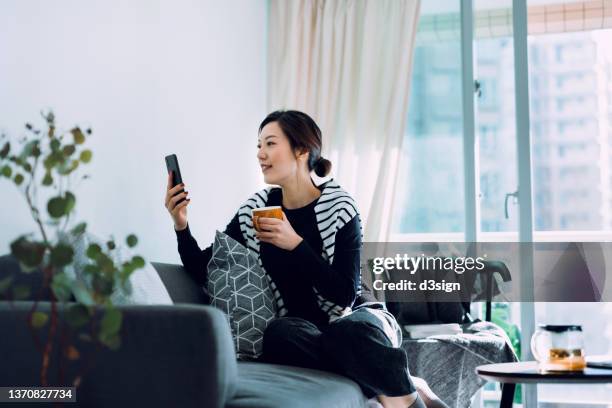 beautiful young asian woman relaxing on sofa with a cup of tea, scrolling on smartphone. enjoying a cozy afternoon at home. lifestyle and technology - asian cinema stock-fotos und bilder