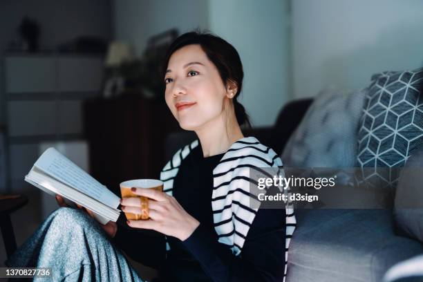 beautiful smiling young asian woman relaxing at home, reading a book with a cup of tea. having a technology-free moment. enjoying a quiet time and relaxing moment at cozy home. healthy lifestyle living - self improvement photos et images de collection