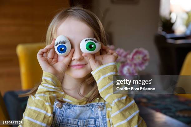 smiling girl (8-9) holding white eggs up to face, with colourful painted eyes and false eyelashes stuck onto them. - eye color stock pictures, royalty-free photos & images