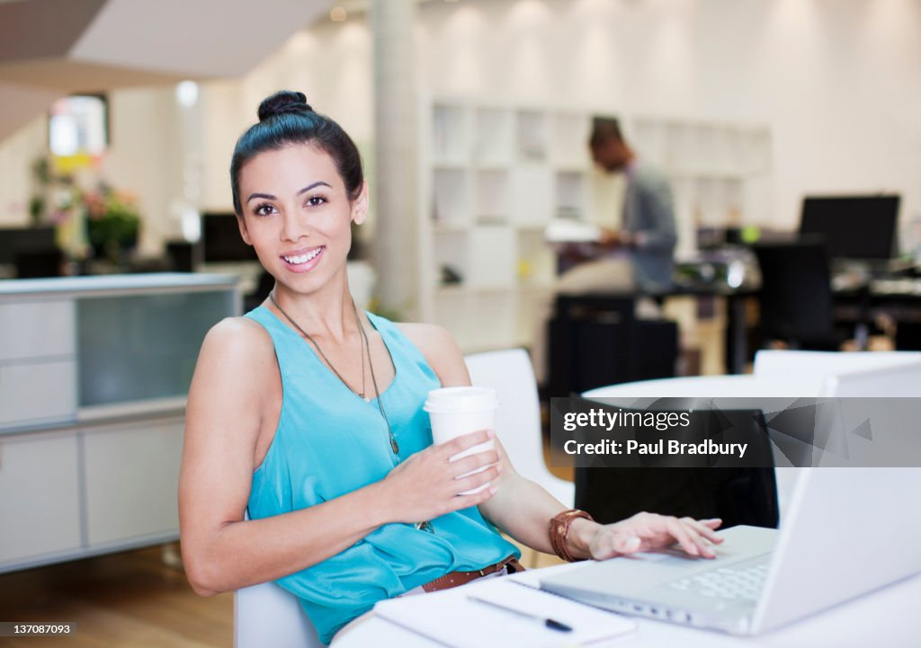 Portrait of smiling businesswoman drinking coffee at laptop in office