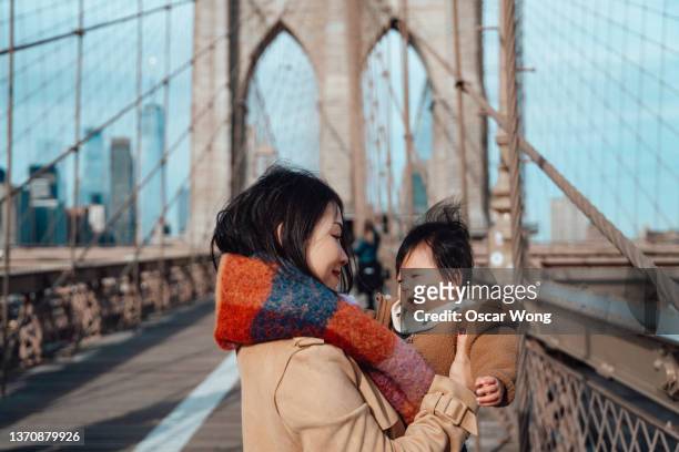 asian mother holding her cheerful baby on brooklyn bridge, new york - new york personas stock-fotos und bilder