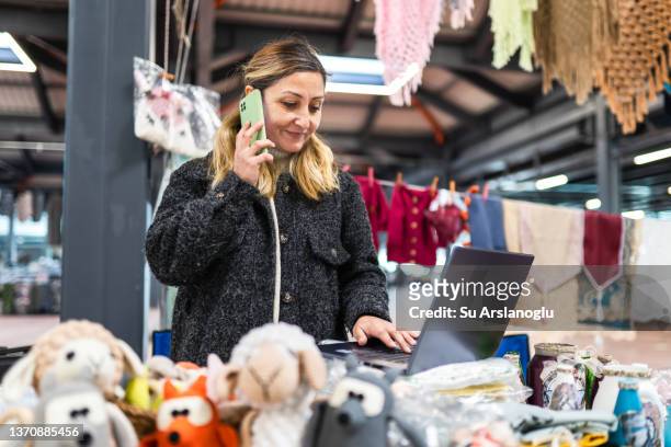 female entrepreneur using laptop and phone in market place in her small business - toy store stock pictures, royalty-free photos & images