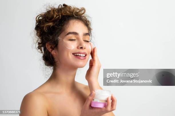 a beautiful woman holds a face cream and applies it on clean skin - ha stock pictures, royalty-free photos & images