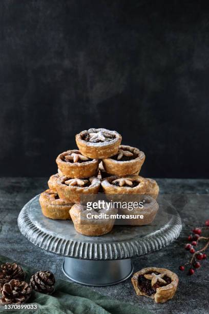 image of batch homemade, mince pies, festive christmas dessert of crispy pastry filled with sweet mincemeat, star shaped pastry top sprinkled with icing sugar, metal cake stand, muslin, pine cones, red berries, black background, focus on foreground - star fruit stockfoto's en -beelden