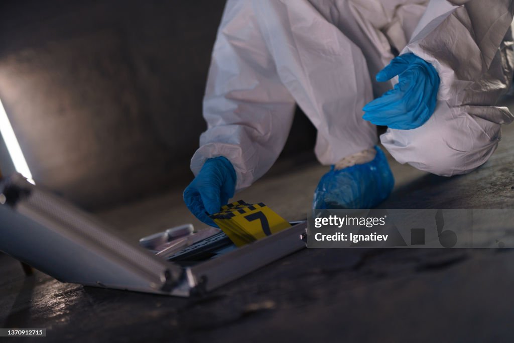 A criminologist at the crime scene, opening a case with criminologist's equipment and takes an evidence number to put it on the flood next to the blood trace