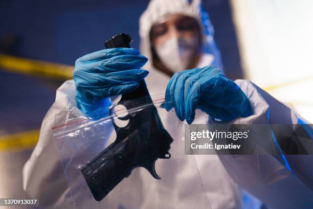 criminólogo con traje protector, máscara y guantes pone una pistola en una bolsa de plástico en la escena del crimen, solo una pistola está en foco - ciencia forense fotografías e imágenes de stock
