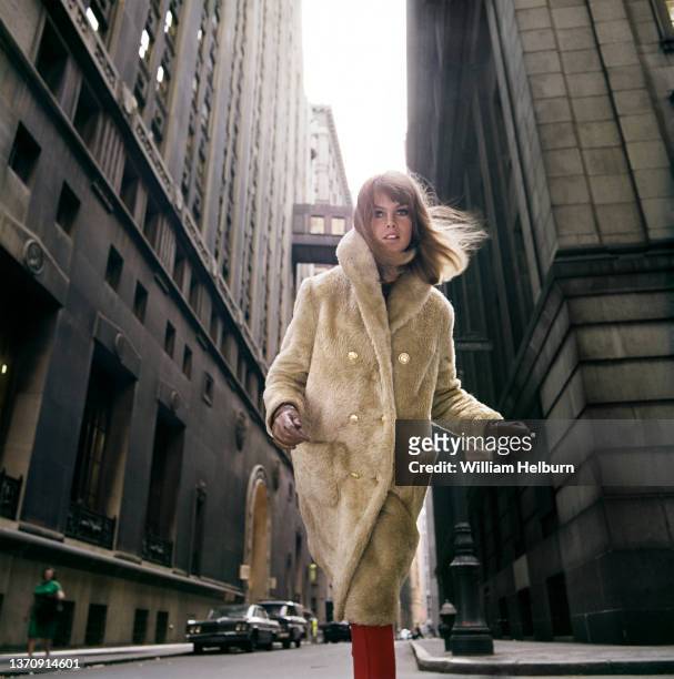 Low-angle view of English model and actress Jean Shrimpton, in a beige fake fur overcoat, brown gloves, and red trousers as she stands in an...